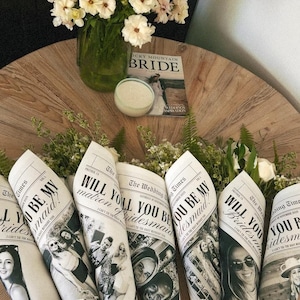 May include: Six bouquets of flowers wrapped in newspaper cones with the text "Will you be my bridesmaid?" printed on them. The bouquets are arranged on a wooden table.