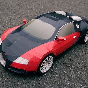 May include: A red and black paper model of a Bugatti Veyron sports car. The model is detailed with a black roof, black wheels, and a red body.