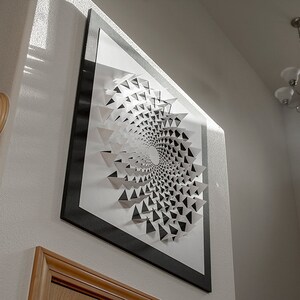 May include: A black and white geometric art print in a black frame. The print features a spiral design made of triangles.