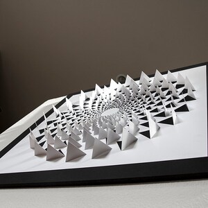 May include: A black framed artwork featuring a white paper sculpture of a spiral pattern made of triangles.