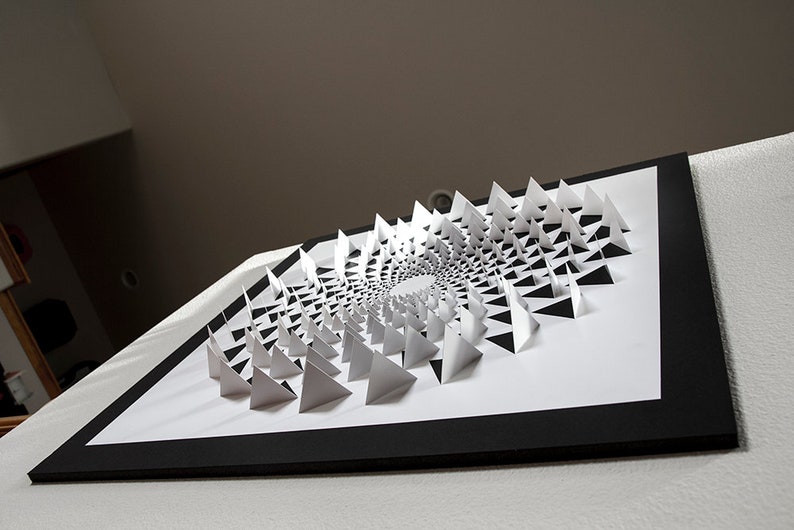 May include: A black framed artwork featuring a white paper sculpture of a spiral pattern made of triangles.