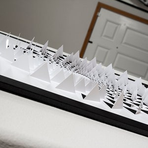 May include: A black framed artwork featuring a white paper sculpture of a spiral pattern made of triangles.