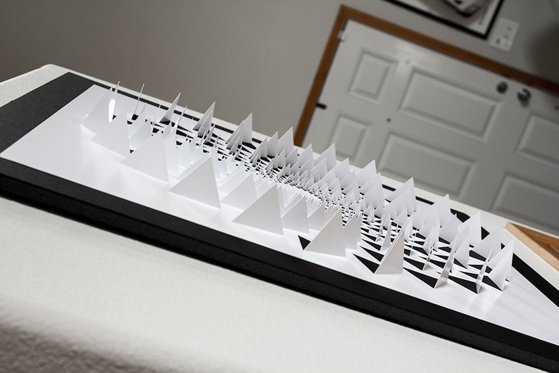 May include: A black framed artwork featuring a white paper sculpture of a spiral pattern made of triangles.