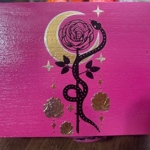 May include: A pink wooden box with a gold crescent moon, rose, and snake design. The design includes gold stars, leaves, and shell shapes. The box has a shiny, smooth finish.