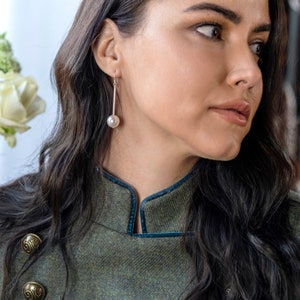 May include: A woman with long dark hair wearing a green tweed jacket with a blue velvet collar and gold buttons. She is wearing a silver earring with a single pearl.