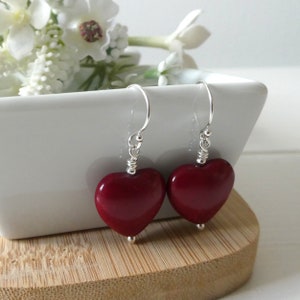 May include: A pair of heart-shaped earrings in a deep red colour. The earrings hang from silver-coloured hooks and have a small silver bead at the base of each heart. The earrings are displayed on a wooden surface.