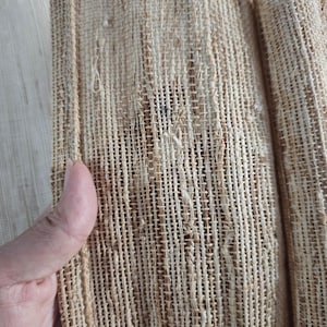 May include: Close-up of a woven fabric with a textured, open weave. The fabric is primarily beige and tan, with visible vertical and horizontal threads creating a grid-like pattern. The material appears to be natural, possibly linen or jute.