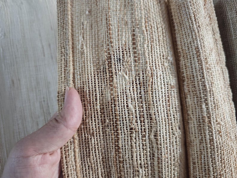 May include: Close-up of a woven fabric with a textured, open weave. The fabric is primarily beige and tan, with visible vertical and horizontal threads creating a grid-like pattern. The material appears to be natural, possibly linen or jute.
