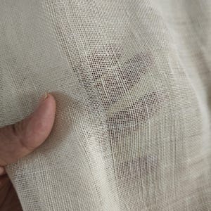 May include: Close-up of a sheer, off-white fabric with a visible grid-like weave. The fabric is held up, allowing light to pass through, revealing a glimpse of a darker surface beneath. The texture appears soft and delicate.