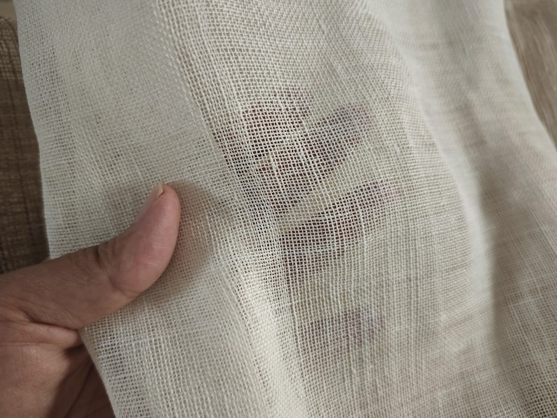 May include: Close-up of a sheer, off-white fabric with a visible grid-like weave. The fabric is held up, allowing light to pass through, revealing a glimpse of a darker surface beneath. The texture appears soft and delicate.