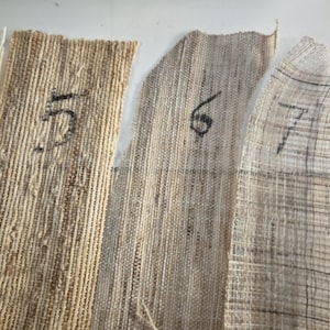 May include: Close-up of several fabric swatches with varying textures and colors, including beige, tan, and off-white. Each swatch is labeled with a number in black ink. The fabrics appear to be natural fibers.
