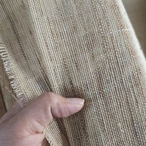 May include: Close-up of a natural burlap fabric. The fabric has a coarse, woven texture with visible fibers and a light brown color. The edge of the fabric is frayed, showing the individual threads. The fabric is being held.