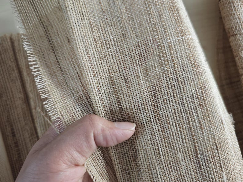 May include: Close-up of a natural burlap fabric. The fabric has a coarse, woven texture with visible fibers and a light brown color. The edge of the fabric is frayed, showing the individual threads. The fabric is being held.