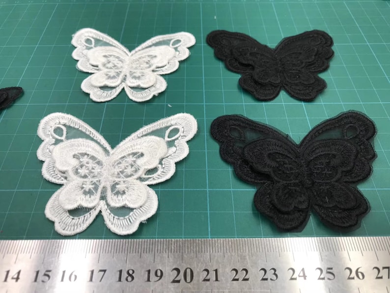 May include: Four butterfly-shaped lace appliques, two white and two black, on a green cutting mat. The appliques are layered and have a delicate, intricate design.