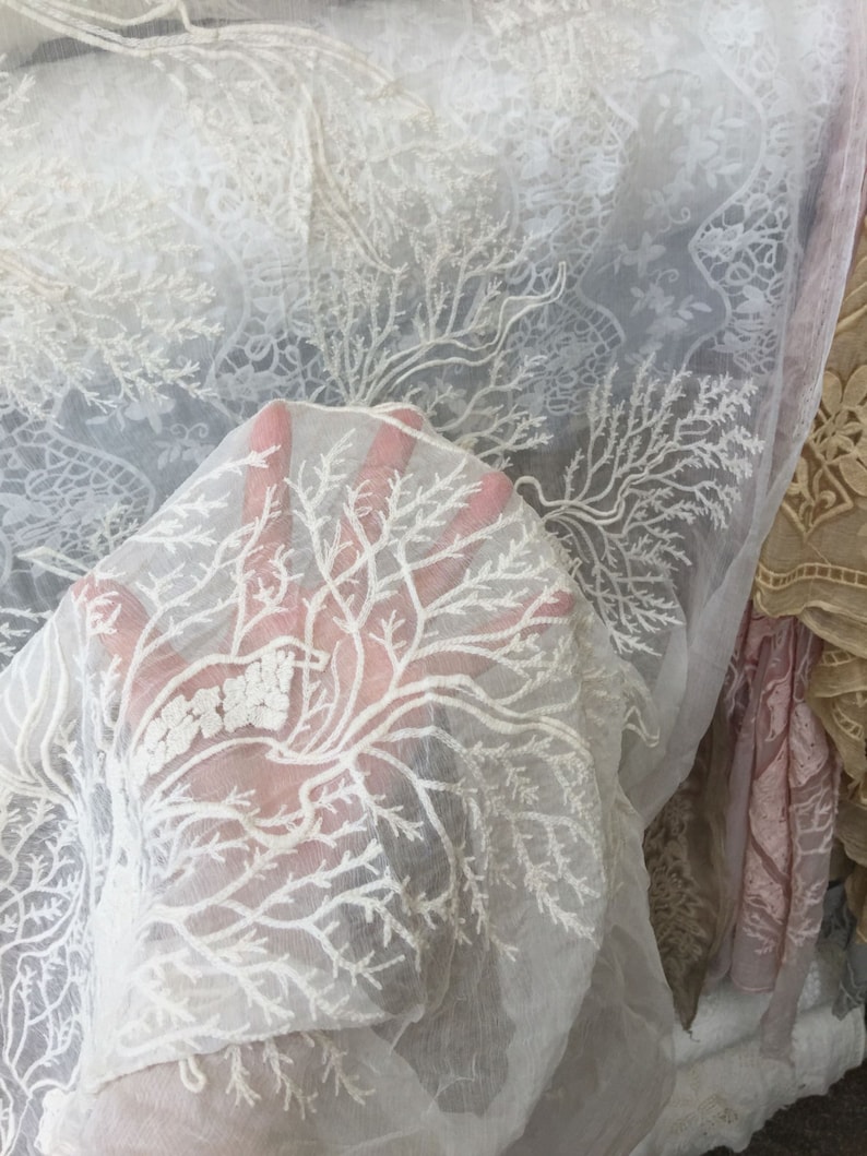Puede incluir: Tela blanca transparente con un delicado patr&oacute;n de encaje floral. El encaje presenta un dise&ntilde;o repetitivo de ramas y hojas.