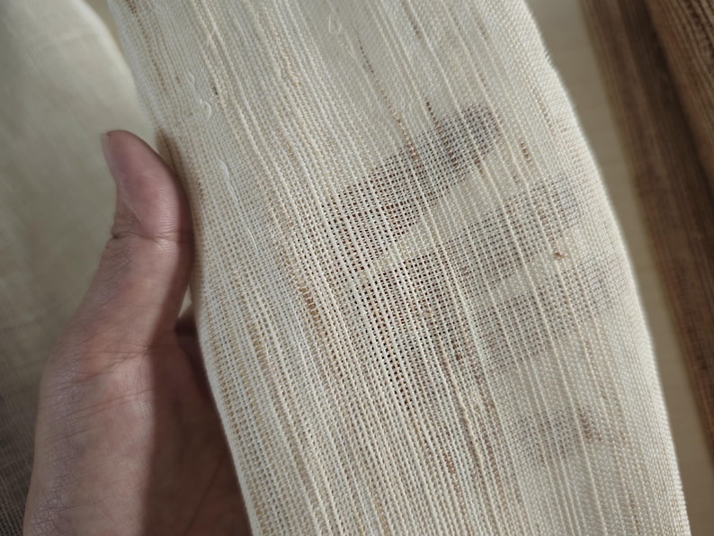 May include: Close-up of a cream-colored, loosely woven fabric. The fabric has a textured appearance with visible threads and small imperfections. The weave is open, allowing some light to pass through. The fabric is being held.