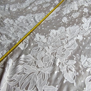 Pode incluir: Tecido de renda floral branca com um padrão delicado. O tecido é transparente e tem uma textura macia e fluida.