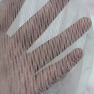 Peut inclure: Gros plan sur un tissu blanc transparent avec un motif de maillage hexagonal fin. Le tissu est maintenu, permettant &agrave; la lumi&egrave;re de passer &agrave; travers, cr&eacute;ant un effet translucide. Le mat&eacute;riau semble d&eacute;licat et l&eacute;ger.