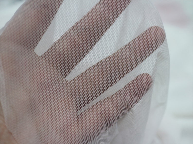 Peut inclure: Gros plan sur un tissu blanc transparent avec un motif de maillage hexagonal fin. Le tissu est maintenu, permettant &agrave; la lumi&egrave;re de passer &agrave; travers, cr&eacute;ant un effet translucide. Le mat&eacute;riau semble d&eacute;licat et l&eacute;ger.