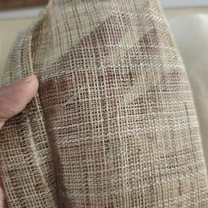 May include: Close-up of a textured, beige fabric with a woven, grid-like pattern. The fabric is held in a hand, revealing its open weave and natural fibers. The background shows a glimpse of a lighter fabric.