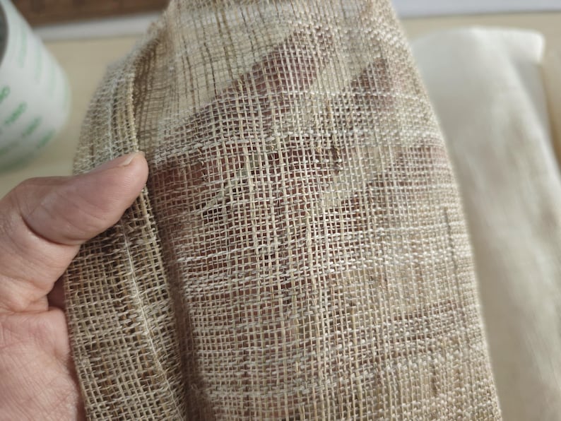 May include: Close-up of a textured, beige fabric with a woven, grid-like pattern. The fabric is held in a hand, revealing its open weave and natural fibers. The background shows a glimpse of a lighter fabric.