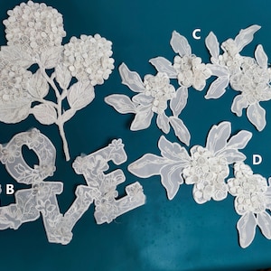May include: Four white lace appliques with floral designs. The appliques are labelled A, B, C, and D. Applique A is a single stem with three flower clusters. Applique B is the word "LOVE" with floral embellishments. Applique C is a vine with three flower clusters. Applique D is a vine with two flower clusters.