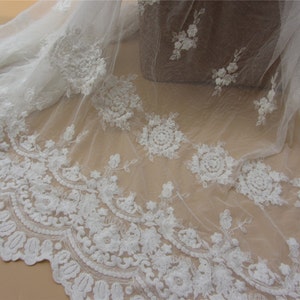 Pu&ograve; includere: Tessuto di tulle bianco ricamato con un delicato motivo floreale. Il tessuto ha una texture morbida e fluida, perfetto per progetti di artigianato e cucito.