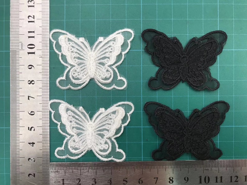 May include: Two pairs of butterfly-shaped fabric appliques. One pair is white and the other is black. The appliques are made of a sheer fabric with a delicate lace design.