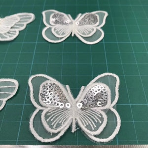 May include: White butterfly-shaped fabric applique with silver sequins. The applique is about 4 inches wide and 3 inches tall.