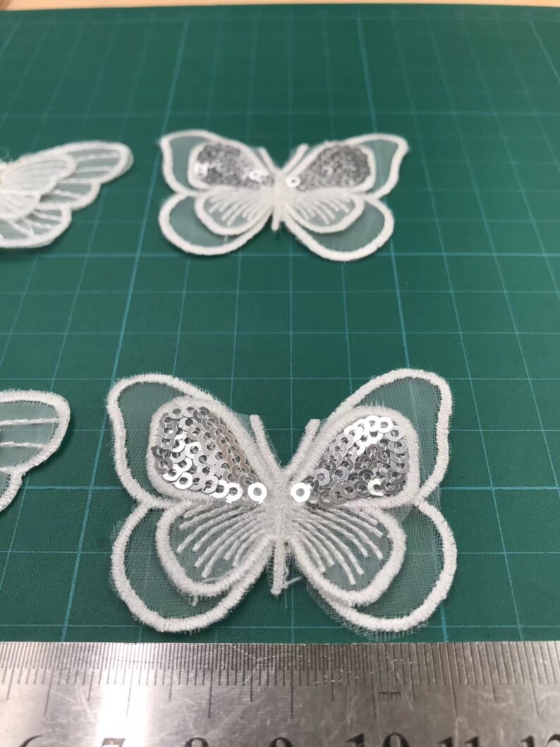May include: White butterfly-shaped fabric applique with silver sequins. The applique is about 4 inches wide and 3 inches tall.