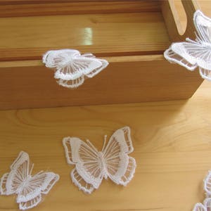 May include: Five white lace butterfly decorations on a wooden surface. The butterflies are delicate and intricate, with a lace pattern.