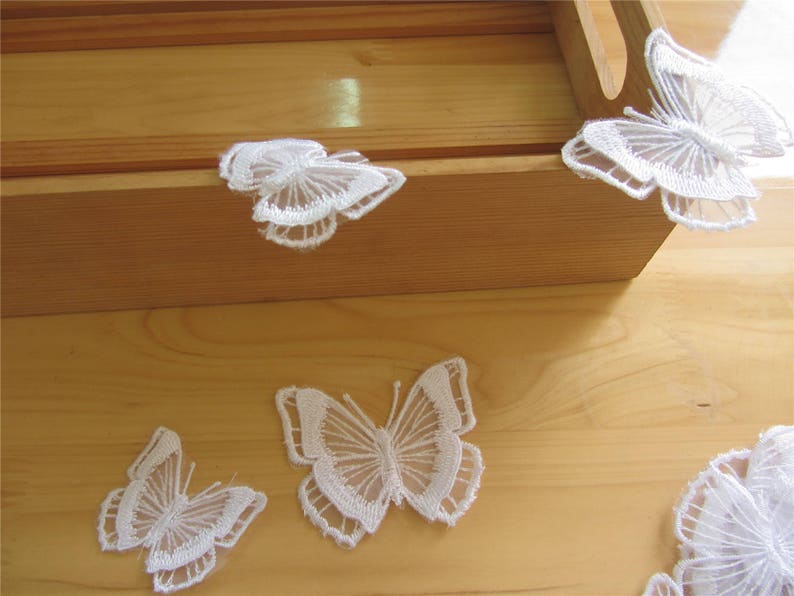 May include: Five white lace butterfly decorations on a wooden surface. The butterflies are delicate and intricate, with a lace pattern.