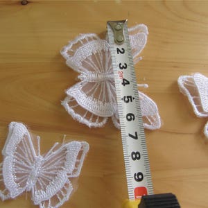 May include: Three white lace butterfly appliques on a wooden surface. A measuring tape is placed next to the largest butterfly, showing a measurement of 6.5 inches.