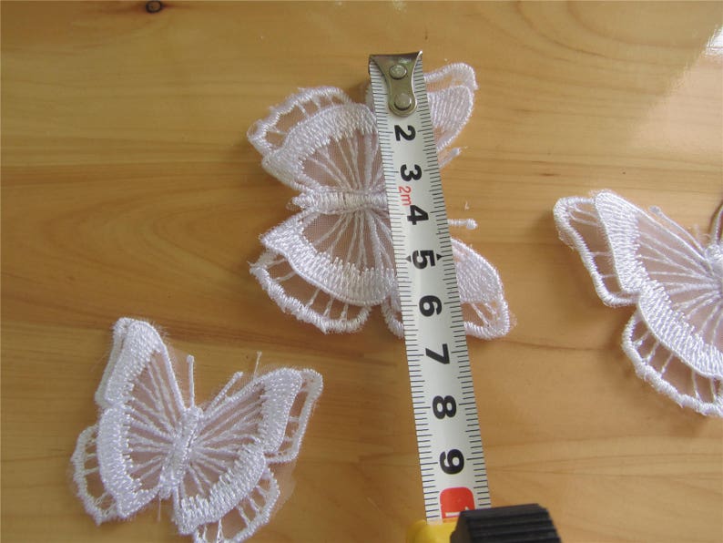 May include: Three white lace butterfly appliques on a wooden surface. A measuring tape is placed next to the largest butterfly, showing a measurement of 6.5 inches.