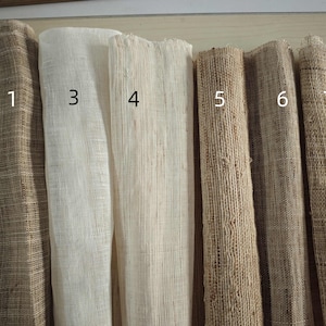 May include: A collection of seven rolls of woven fabric in various neutral tones. The fabrics range in color from light beige to dark brown, with visible textures and varying weaves. The rolls are numbered 1 through 7.