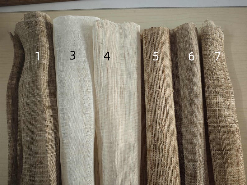 May include: A collection of seven rolls of woven fabric in various neutral tones. The fabrics range in color from light beige to dark brown, with visible textures and varying weaves. The rolls are numbered 1 through 7.