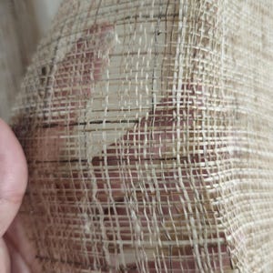 May include: Close-up of a woven fabric with a natural, beige color. The weave is open, creating a grid-like pattern with visible gaps. The texture appears rough and organic, suggesting a handmade or natural fiber material.