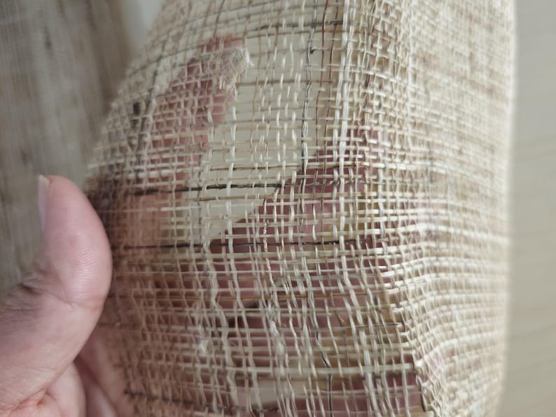 May include: Close-up of a woven fabric with a natural, beige color. The weave is open, creating a grid-like pattern with visible gaps. The texture appears rough and organic, suggesting a handmade or natural fiber material.