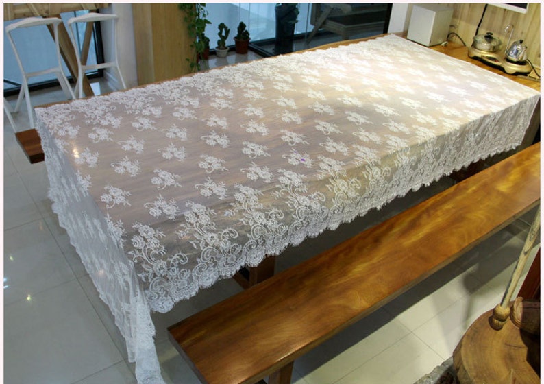 K&ouml;nnte beinhalten: Wei&szlig;e Tischdecke aus Spitze mit Blumenmuster, &uuml;ber einen Holztisch und eine Holzbank gelegt.