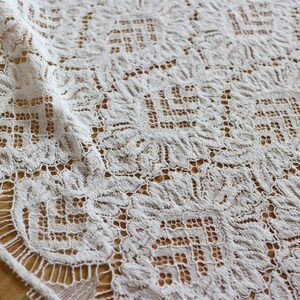 May include: Close-up of a white lace fabric with a floral and geometric pattern. The delicate lace is draped, revealing intricate details and textures. The fabric's edges are finished with a scalloped trim, adding to its elegance. The background is a light brown surface.