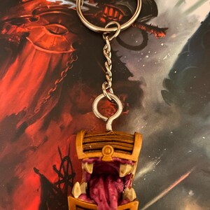 Könnte beinhalten: Ein Schlüsselanhänger in Form einer Holzkiste mit einem zahnigen, offenen Mund und einer heraussteckenden rosa Zunge. Der Schlüsselanhänger ist an einer silbernen Kette und einem Ring befestigt.