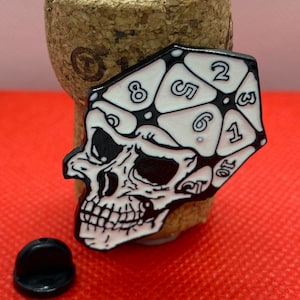 May include: Black and white enamel pin featuring a skull with a white d20 die on top. The die shows the numbers 1, 2, 3, 4, 5, 6, 7, 8, 9, 10, 11, 12, 13, 14, 15, 16, 17, 18, 19, and 20.