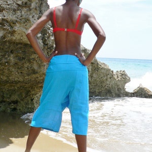 May include: A person wearing a bright blue pair of harem pants and a red bikini top, standing on a sandy beach with a rocky cliff in the background.