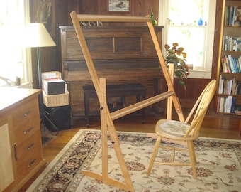 May include: A wooden tapestry frame with adjustable height and a folding base. The frame is set up in a room with a rug, a chair, and a piano.