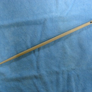 May include: A long, thin, wooden dowel rod with a small hole drilled at one end. The rod is light brown in color and is lying on a blue fabric surface.