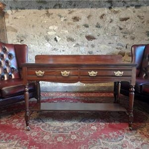 May include: A mahogany console table with two drawers, each with gold-coloured crown-shaped handles. The table has a lower shelf and stands on turned legs with small castors. It is set against a stone wall and a patterned red rug.