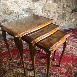 May include: A set of three nested tables crafted from brown wood with a polished finish. Each rectangular table features a smooth top surface with a visible wood grain pattern, carved decorative edges, and slender cabriole legs with ball feet.