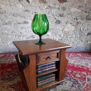 May include: A wooden magazine rack with a drawer and two shelves. The rack is on a red and cream patterned rug. A green glass vase sits on top of the rack.