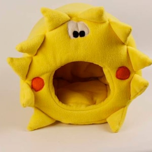 Puede incluir: Una cama de mascota de felpa amarilla con forma de sol con detalles naranjas y una gran abertura para que una mascota pueda entrar.