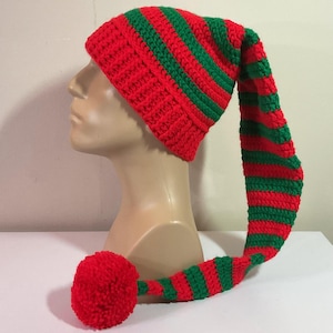 May include: A red and green striped elf hat with a long, pointed end and a red pom-pom. The hat is made of a knitted material and is displayed on a mannequin head. The hat is a festive accessory.